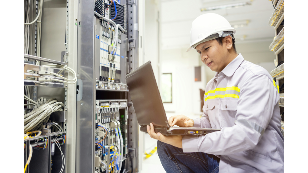Electrical engineer in safety gear inspecting server racks while working on a laptop, surrounded by organized cables and hardware.