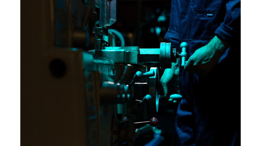 Close-up of a technician operating a lathe or precision industrial machine, illuminated by blue-tinted lighting in a dim workshop.