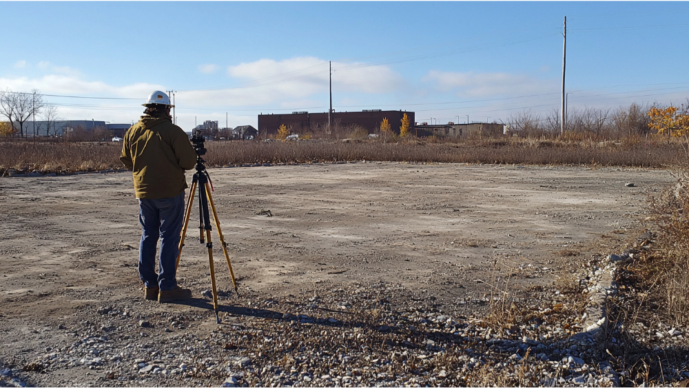 Surveyor standing on an open lot using a tripod and surveying instrument to measure land boundaries and prepare for construction.