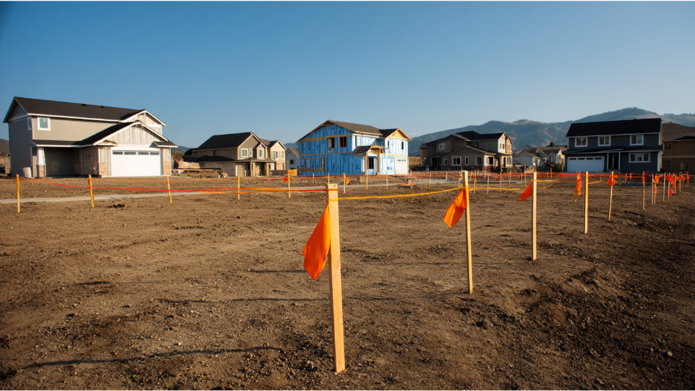 Construction site with orange boundary flags outlining new home lots in a suburban development under clear skies.