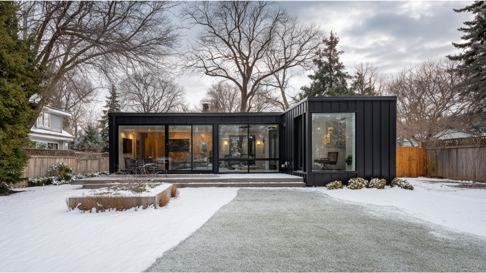 Flat-roof ADU with black siding and large floor-to-ceiling windows, set in a snow-covered backyard with trees and fencing.