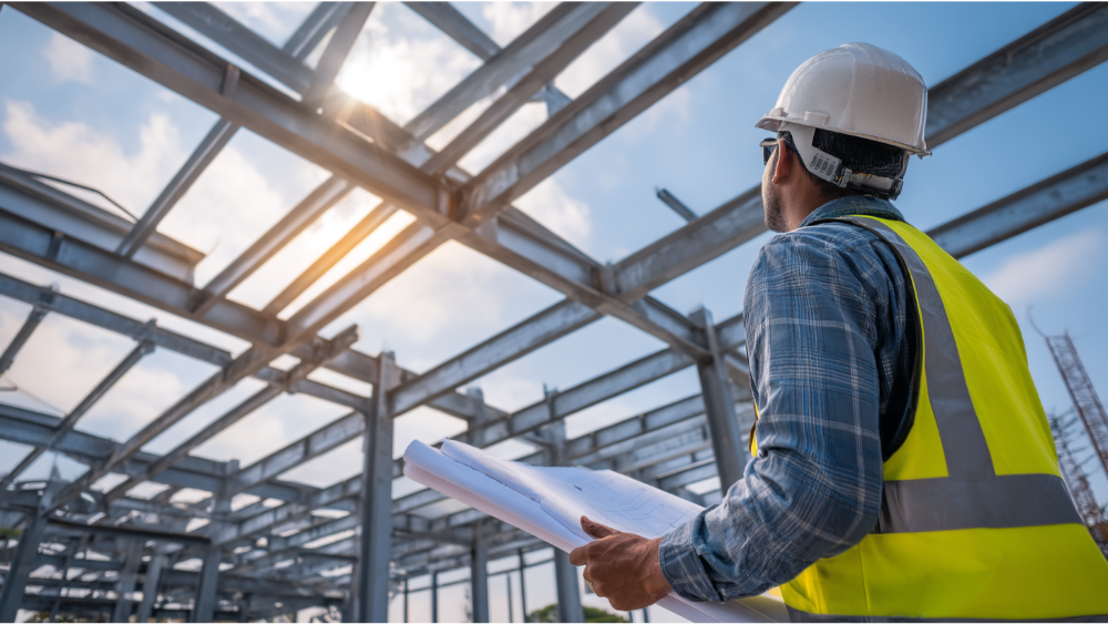 A professional in a hard hat and high-visibility vest looks up at a steel building framework while holding blueprints under a partly cloudy sky.