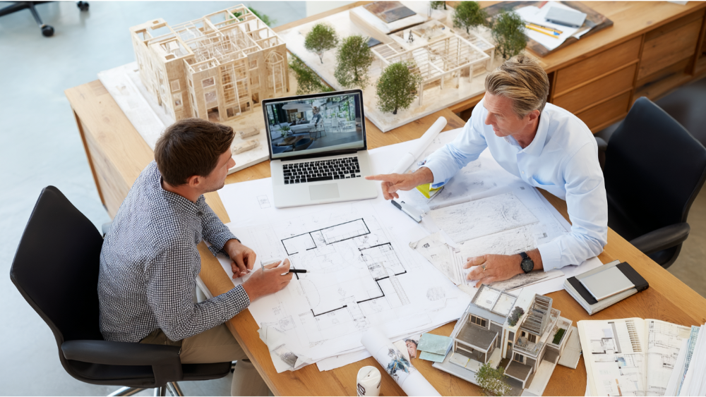 Two professionals engaged in a design discussion over residential floor plans and architectural models.