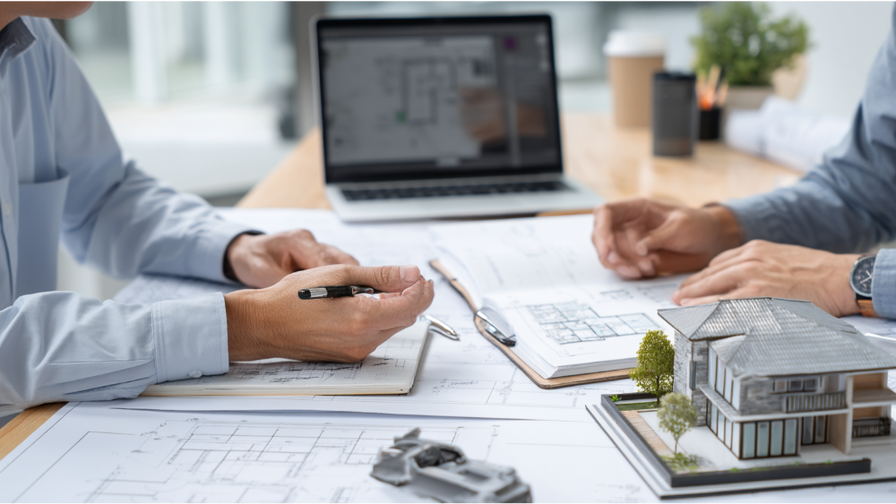 Architects collaborating at a desk covered with blueprints, a laptop, and a miniature house model.