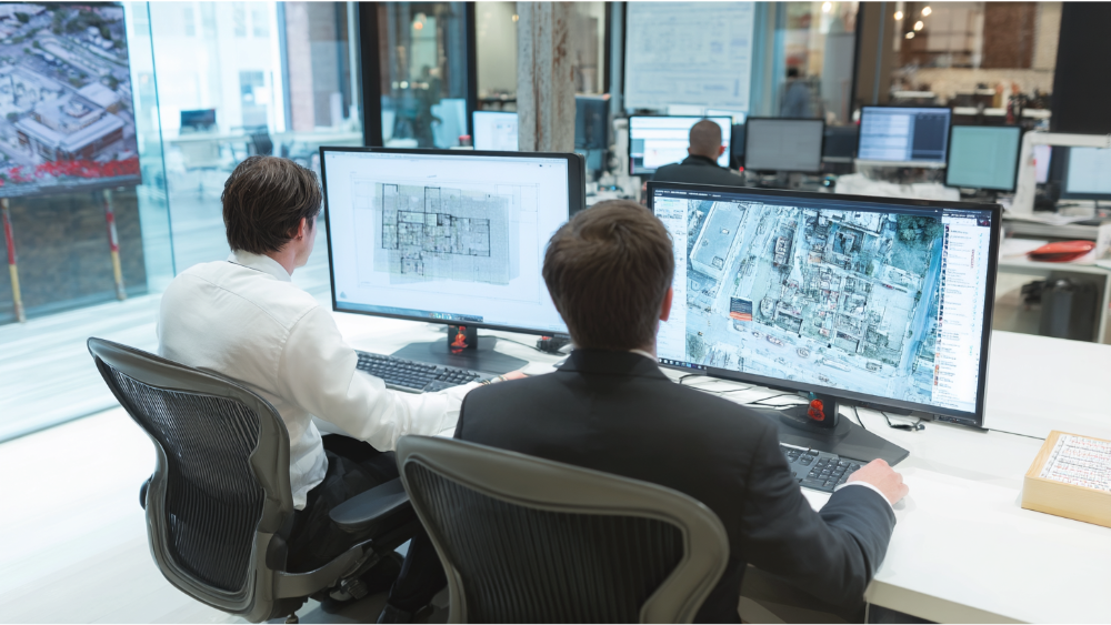 Two professionals reviewing architectural and aerial site plans on dual monitors in a modern office.