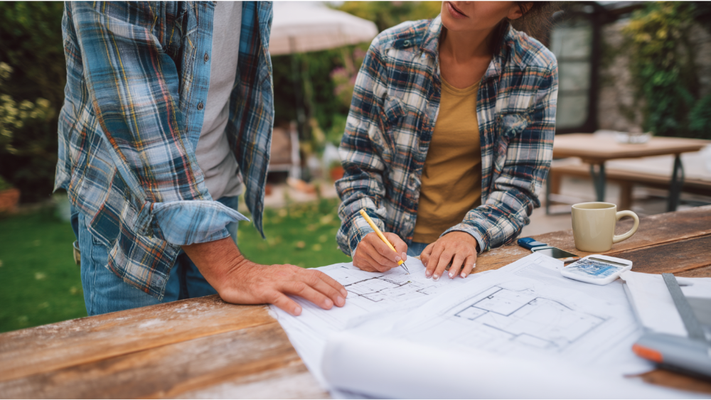 Two homeowners reviewing lot line adjustment plans on a wooden table outdoors, with printed blueprints and measuring tools.