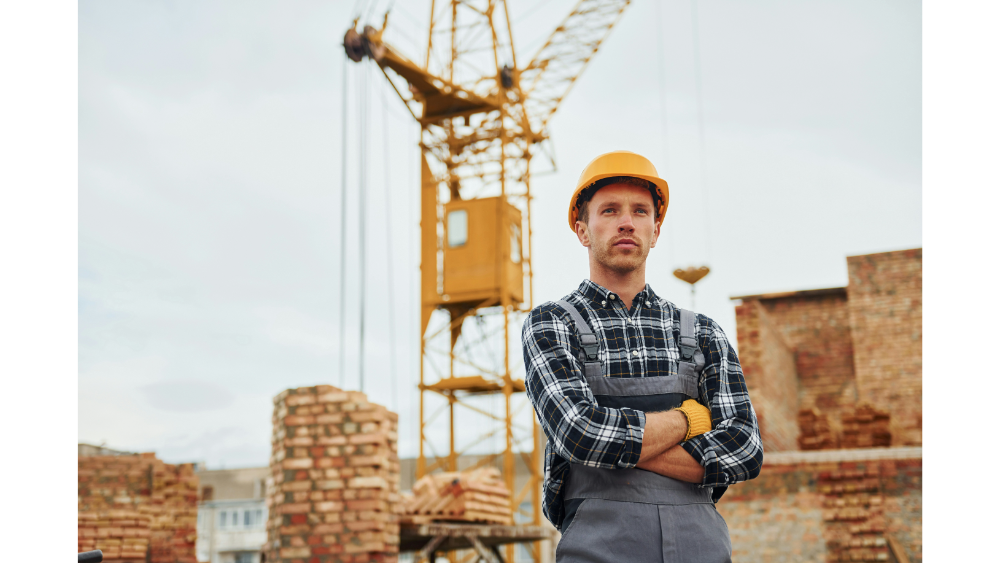 A man wearing a hard hat and plaid shirt stands with folded arms in front of a large crane and partially built brick structures.