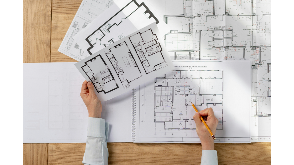 A person analyzing various architectural blueprints and layout drawings, marking one of the plans with a pencil at a design workspace.