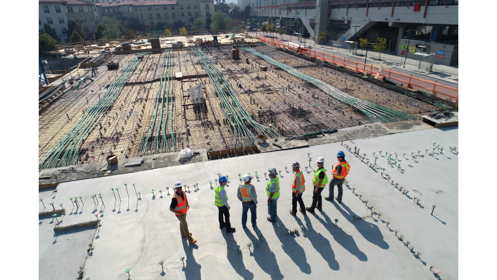 A group of workers and supervisors wearing safety gear standing on a large concrete slab surrounded by steel rebar and utility conduits.