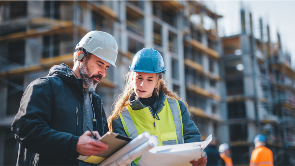Construction workers in hard hats and safety vests reviewing blueprints at an active building site.