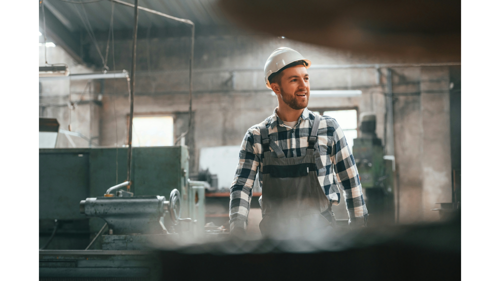 Smiling factory worker in a plaid shirt and hard hat standing in a workshop with heavy machinery in the background